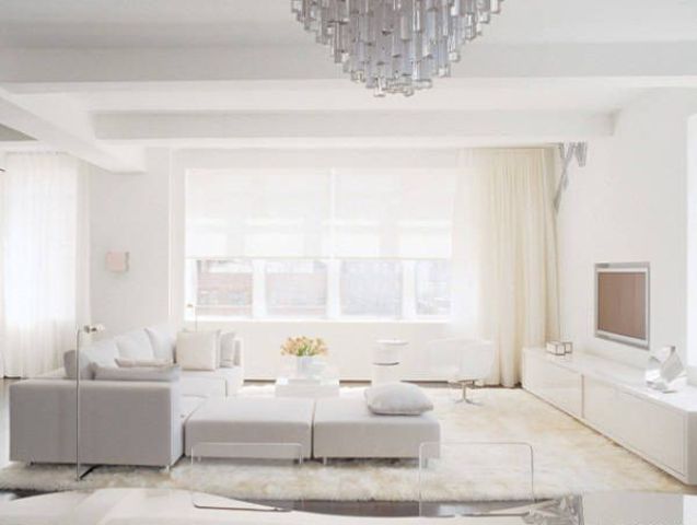 A modern white living room with a neutral sofa, a couple of ottomans and layered rugs, a sleek storage unit and a crystal chandelier