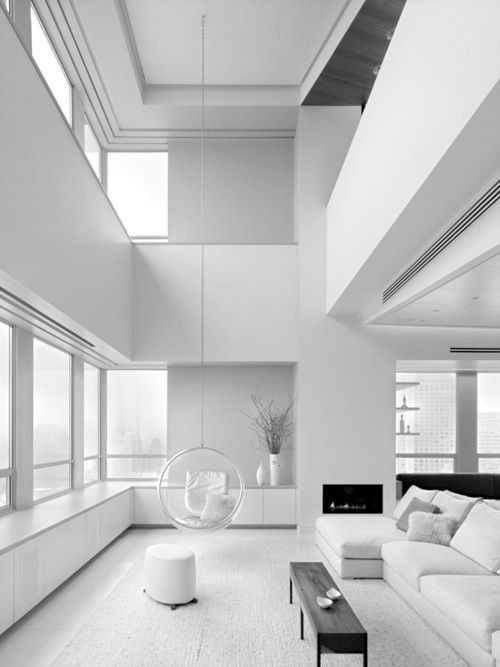A minimalist living room with double height ceilings, a built in fireplace, a low black coffee table and a pendant clear chair