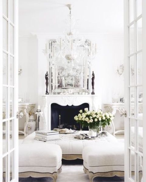 A white vintage living room with a chic non working fireplace, a large low ottoman and poufs, a crystal chandelier and a large mirror is amazing