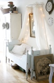 a neutral shabby chic kid’s room with a pastel blue bed, white furniture, a canopy with lights and some vintage decor