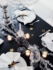 a black and white NYE party tablescape with white porcelain, glasses, candles, silver branches and small stars on the table