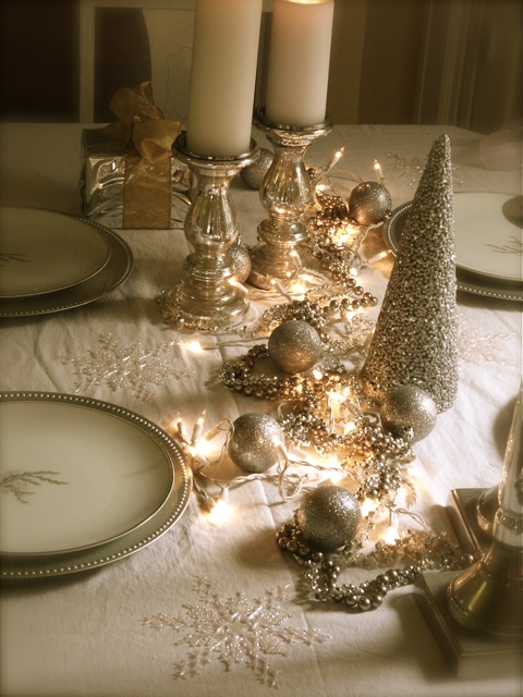 A gold and white NYE tablescape with gold rimmed glasses, gold glitter ornaments and a cone shaped tree, candleholders with pillar candles