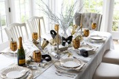 a white NYE tablescape with a silver table runner, white plates, gold glasses, gold candles and masks is an elegant idea