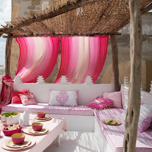 A bold Mediterranean terrace with a built in bench with pink upholstery and hot pink curtains, a small roof over the bench and a low coffee table