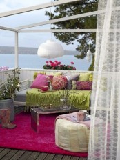 a colorful terrace with a sea view, with a green sofa with pink and green pillows, a low coffee table and a green pouf, pendant lamps and potted blooms