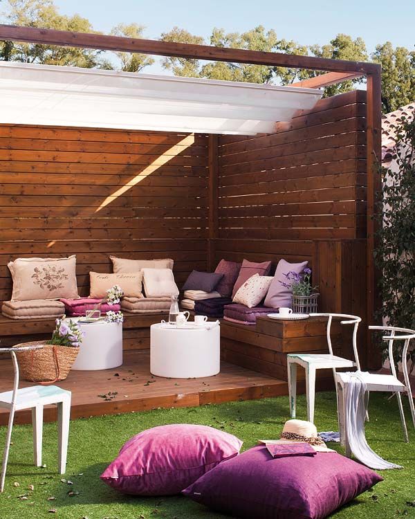 A lovely feminine terrace with stained wood around, with a built in bench, pink, purple and pastel upholstery, white round coffee tables and white chairs, purple pillows