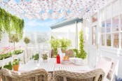 a bright feminine terrace with white planked tables, woven chairs, floral upholstery and a roof and potted plants and blooms around