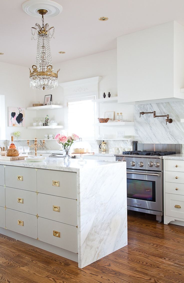 A white glam kitchen with marble countertops, gold handles and knobs, a vintage crystal chandelier over the space