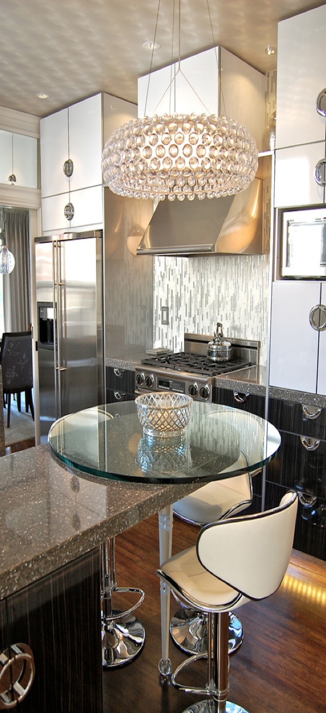 A glam kitchen with dark cabinetry, shiny silver handles, a large crystal chandelier and a glass countertop
