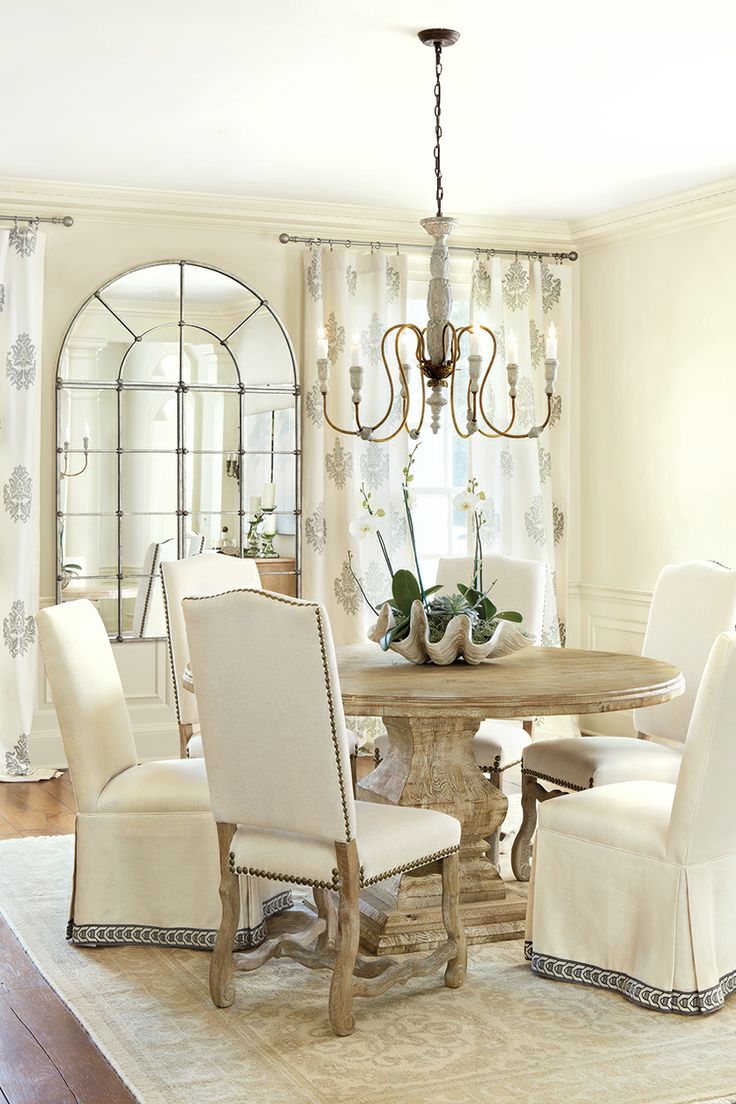 A warm colored vintage dining room with tan walls, a round stained table and chairs covered with fabric, with a vintage chandelier and a French window