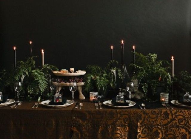 A Halloween tablescape with a rust printed tablecloth, black and white candles, lots of greenery and refined plates and glasses