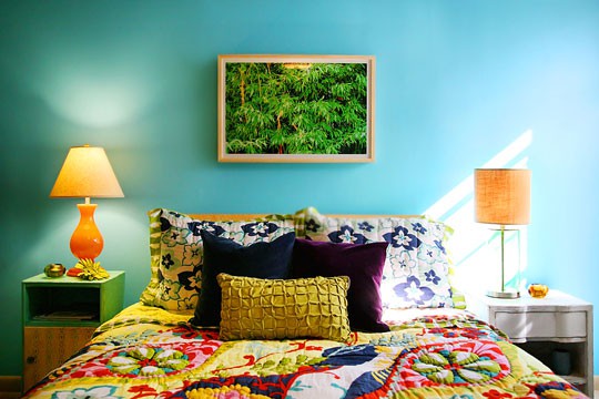 a colorful bedroom with turquoise walls, a bed with colorful bedding, mismatching nightstands, table lamps and a bold artwork and bright pillows