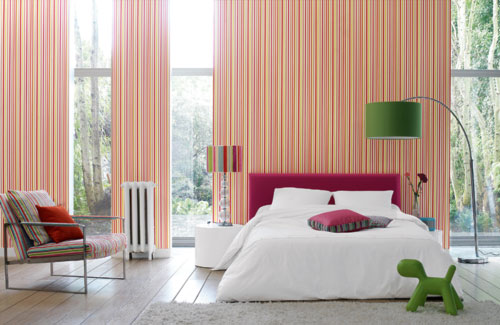 a bright bedroom with colorful striped walls, an upholstered fuchsia bed, a striped chair with a cfuchsia pillow, a green floor lamp and a green toy is fun and bold