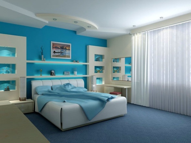 A turquoise and white bedroom with built in storage units, a white upholstered bed and turquoise bedding, a mirror and a desk and white curtains