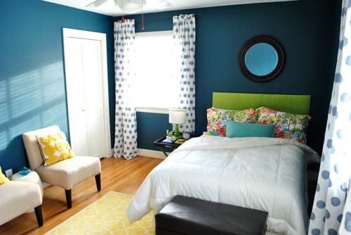 a navy bedroom with a bright grene bed, colorful pillows and a yellow printed rug, bright pillows on the neutral chairs