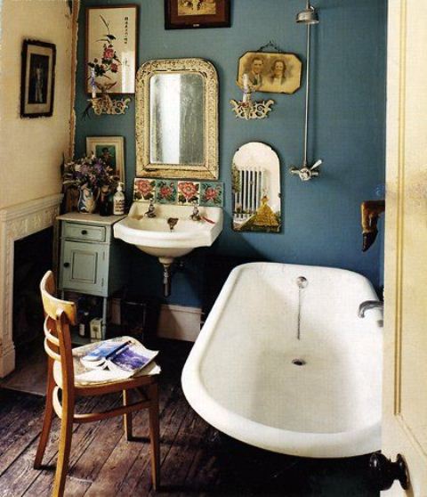 A vintage bathroom with blue walls, a free standing tub, a gallery wall and artworks plus vintage furniture