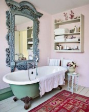 a pink bathroom with a green clawfoot tub, boho rugs, a large ornate mirror in a blue frame and a shelf