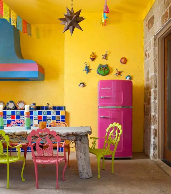 a colorful space with yellow walls, a colorful tile backsplash, a whitewashed rough wood table, yellow and pink chairs and colorful decorations plus a pink fridge
