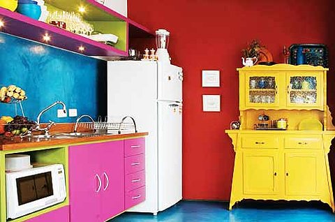 a crazy colorful kitchen with a bold blue and bold red wall, a bright yellow buffet, hot pink cabinets and niches in green, with lights