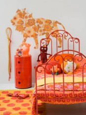 a whimsical neutral girl’s bedroom with a red metal bed with yellow and orange bedding, a red nightstand, a yellow floral mural, bright toys