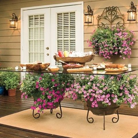a spring patio with a creative table placed on blooming planters, with a blooming planter on the wall, some potted greenery and a glass table