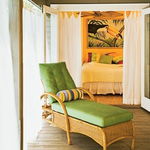bright bedding and printed pillows, a statement artwork, a wicker lounger with green upholstery for a tropical feel