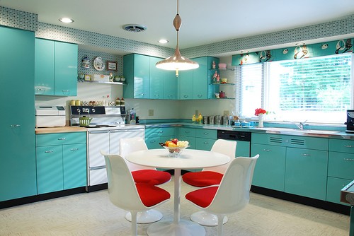 a bright turquoise kitchen with sleek retro cabinetry, a white table and white and red dining chairs and lights and lamps