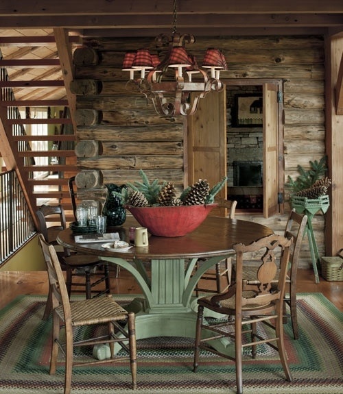 A rustic chalet dining room with a dark stained round table, matching chairs, a red lampshade chandelier and a bowl with pinecones and evergreens