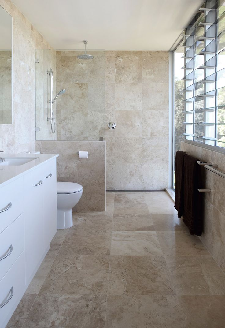 A tan colored stone tile bathroom with a white vanity and white appliances plus a glazed wall