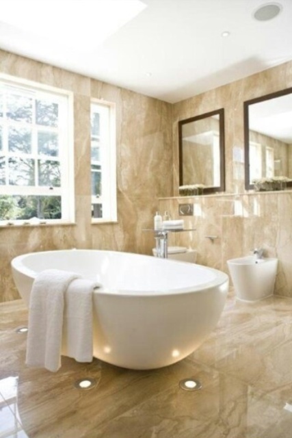 a tan abthroom clad with marble tiles all over, white applainces, mirrors and much natural light coming from outside
