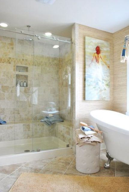 A tan colored bathroom clad with tiles, with neutral wallpaper, a vintage bathtub and a shower space plus a bold artwork