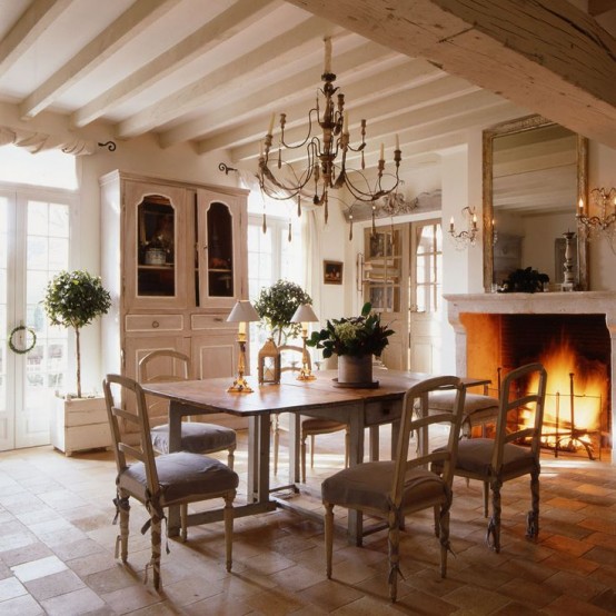 an elegant neutral Provence dining room with a large fireplace, a folding table and vintage chairs, a buffet, some potted plants and chandeliers