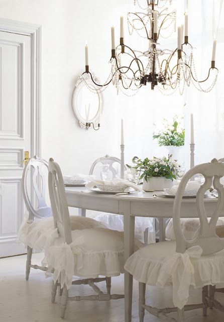 a pretty white Provence dining space with a white table and vintage chairs with cushions, a chandelier, an oval mirror, greenery and blooms