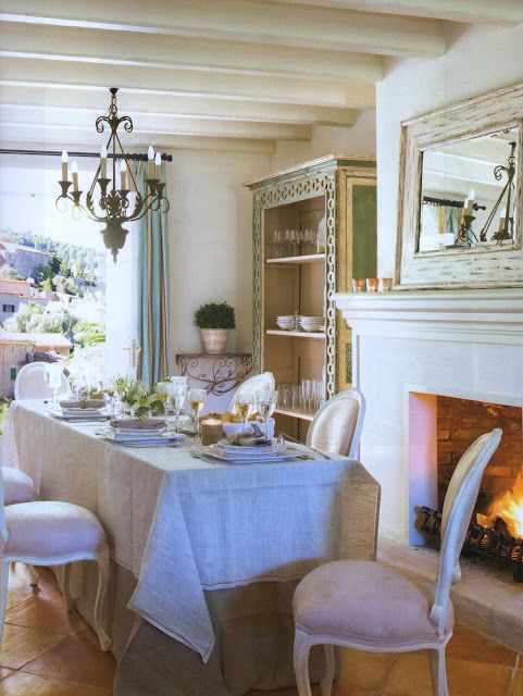 a beautiful neutral Provence dining space with a fireplace, a table with a linen tablecloth, vintage chairs, a chandelier, a buffet and a large mirror