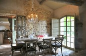 a Provence dining room with stone walls, a window with arched doors, a stained table and chairs, a vintage chandelier and a tiled floor