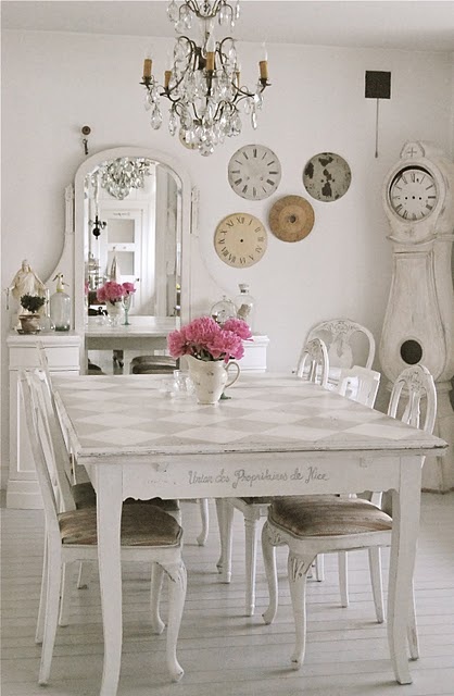 a white vintage Provence-inspired dining room with a vanity with a mirror, a grandfather's clock, a white table and refined chairs, a chandelier and some decorative plates