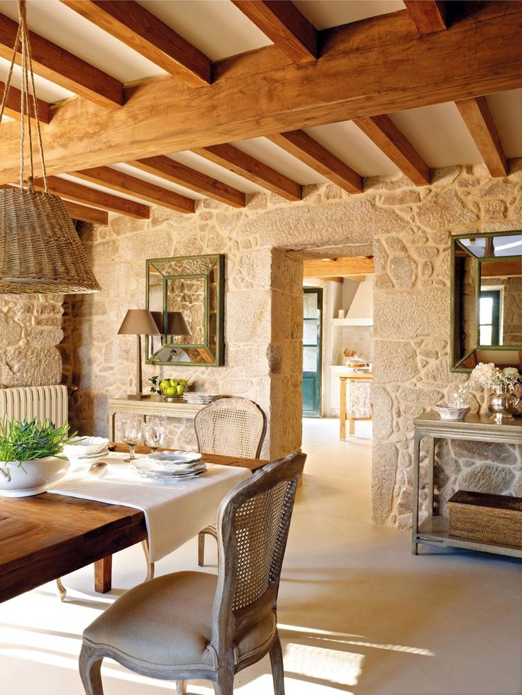 A rustic Provence dining room with stone walls and a wooden ceiling with beams, a stained table and vintage chairs, console tables, wicker lamps and mirrors