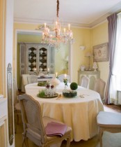 a French chic dining room with pastel yellow walls, a corner cabinet, an oval table and vintage chairs, a crystal chandelier and greenery topiaries