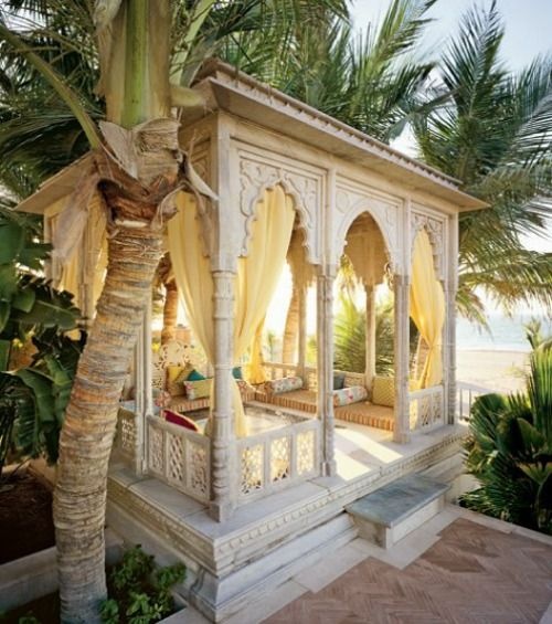 a white Moroccan cabana of carved wood with a long upholstered bench and bright pillows
