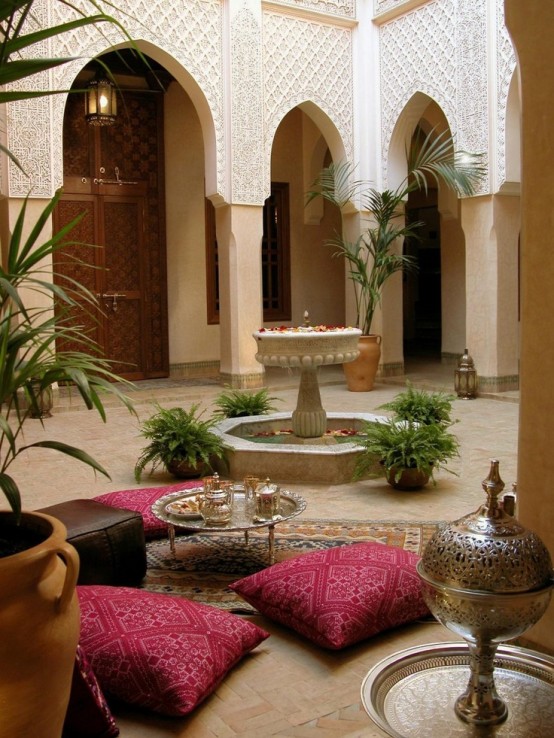 a cozy Moroccan patio with pink pillows, a fountain and potted greenery, low furniture and ottomans