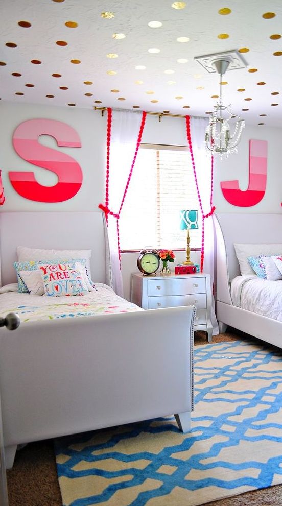 a colorful shared girls' bedroom with creamy upholstered beds, colorful bedding, a bright rug, pink to red ombre monograms, a gold polka dot ceiling for a touch of fun