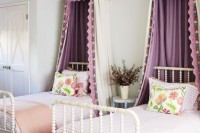 a chic and stylish shared girls’ bedroom with white vintage beds, lilac and peachy bedding, purple canopies, a built-in wardrobe and a woven basket for storage