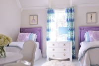 a refined shared girls’ bedroom with purple upholstered beds, purple, white and blue bedding, a chic white dresser and a white table plus a chair, teal printed curtains