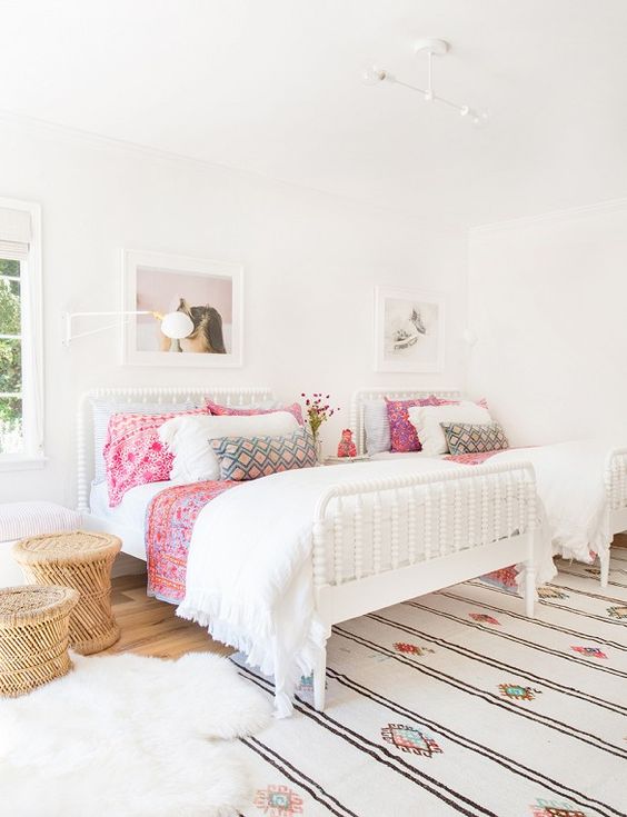 a chic neutral boho shared girls' bedroom with white wooden beds, a bright printed rug, pink bedding, wooden side tables, faux fur and blooms