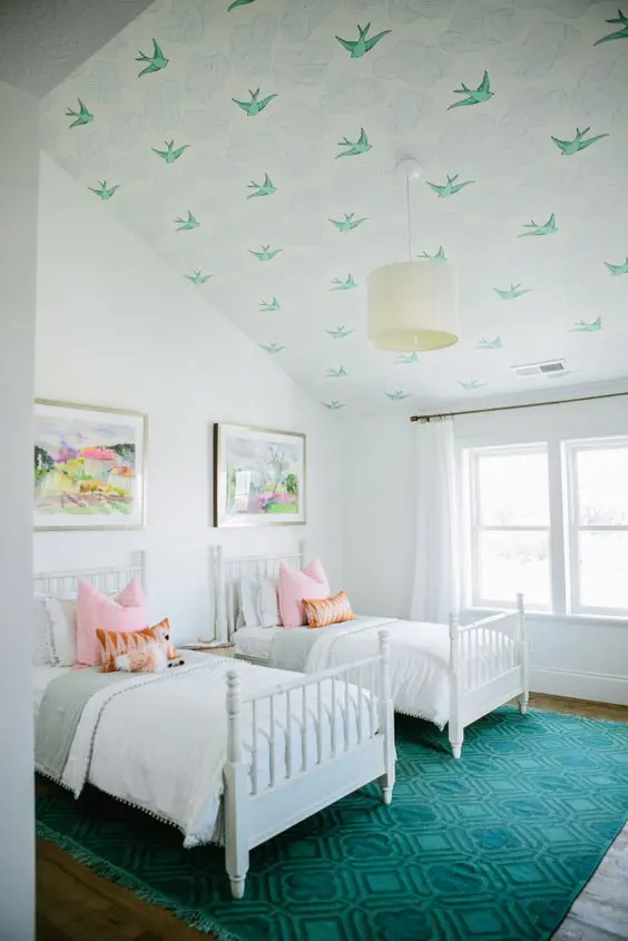 A dreamy white shared attic bedroom with a bird printed ceiling, white vintage beds, neutral and pastel bedding, a teal rug and a bright artworks is cool