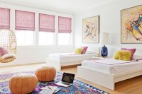 a chic shared girls’ bedroom with a series of windows with pink curtains, contemporary beds, bright artworks, a colorful rug and orange leather poufs