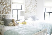 a lovely pastel shared girl bedroom with an accent botanical wall, white wooden beds, blue and grey bedding, a bright nightstand and a table lamp