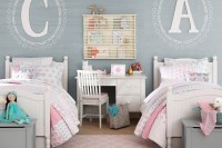 a shared girls’ bedroom with a grey accent wall with monograms, white vintage beds and grey chests for storage, pink bedding, a white desk with a chair that doubles as a nightstand