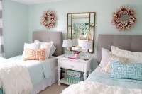 a stylish pastel girls’ bedroom with light blue walls, grey upholstered beds, blue and white bedding and pillows, fabric wreaths and a framed mirror on the nightstand