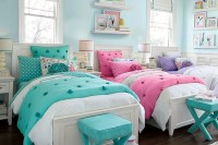 a pastel shared girls’ bedroom with a light blue accent wall, three vintage neutral beds, purple, pink and turquoise bedding, turquoise stools, bookshelves on the walls